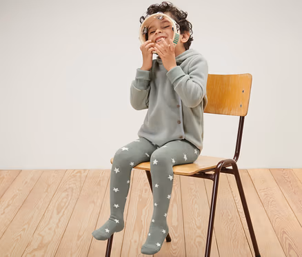 Ein Junge sitzt auf einem Stuhl und spielt mit einem Tamburin. Er trägt eine blaue und grüne Kinder-Thermostrumpfhose.
