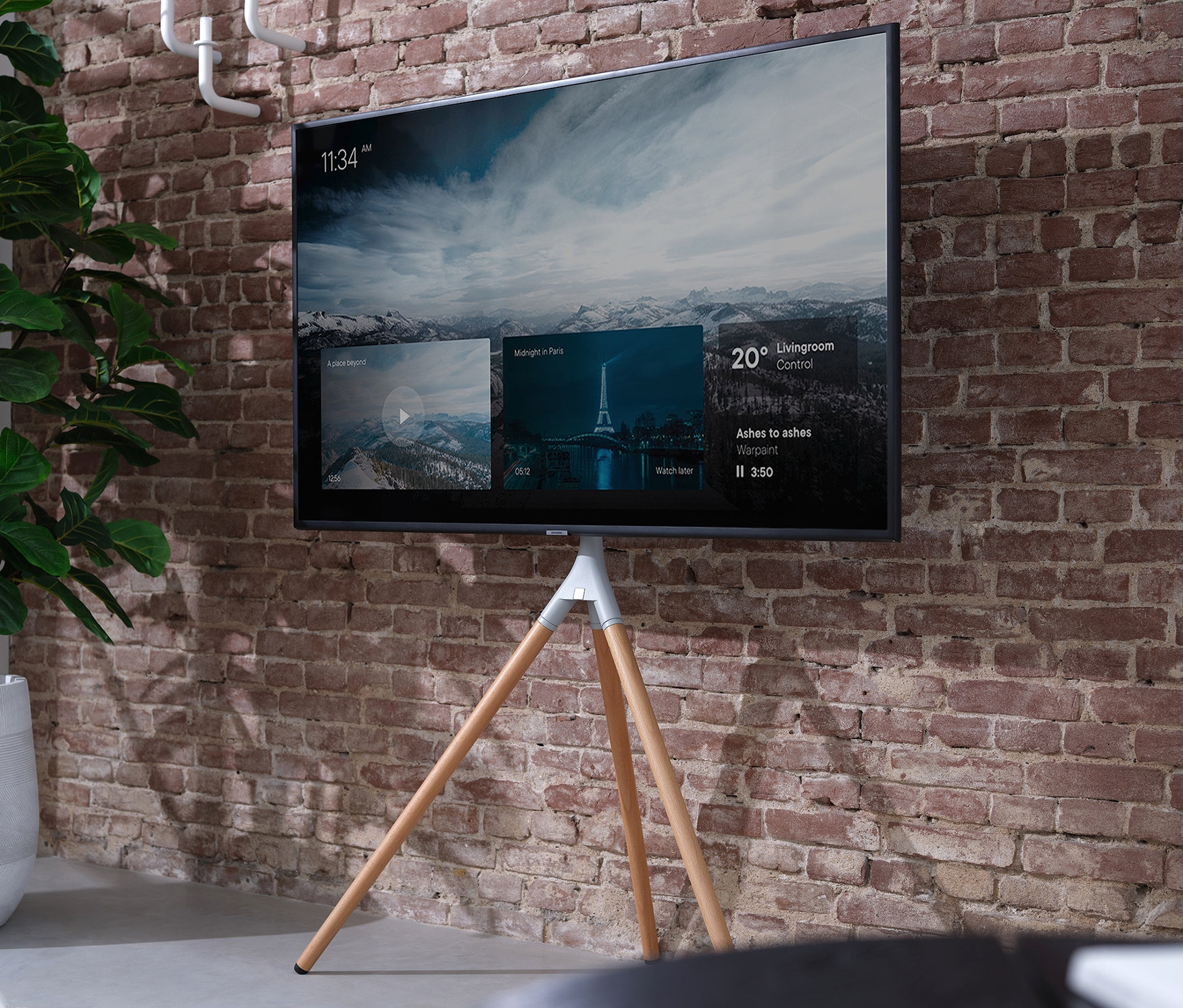 Ein Fernseher steht auf einer ONE FOR ALL TRIPOD-TV-Halterung »WM7472« in Eiche und Silbergrau vor einer Ziegelwand. Neben dem Fernseher befindet sich eine Pflanze in einem Topf.