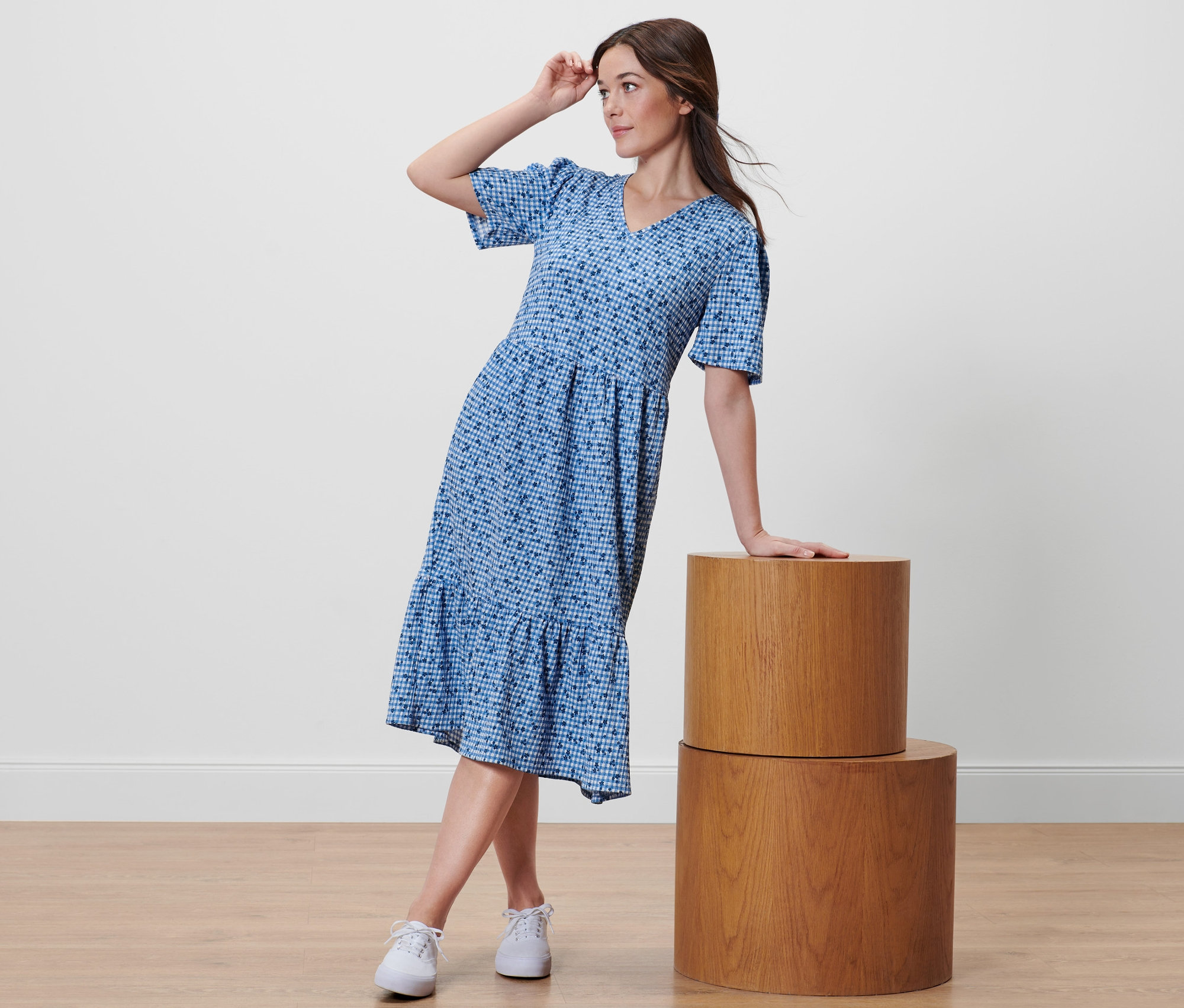 Frau posiert in blauem Kleid mit Alloverprint und weißen Turnschuhen, während sie sich an zwei Holzzylindern anlehnt.