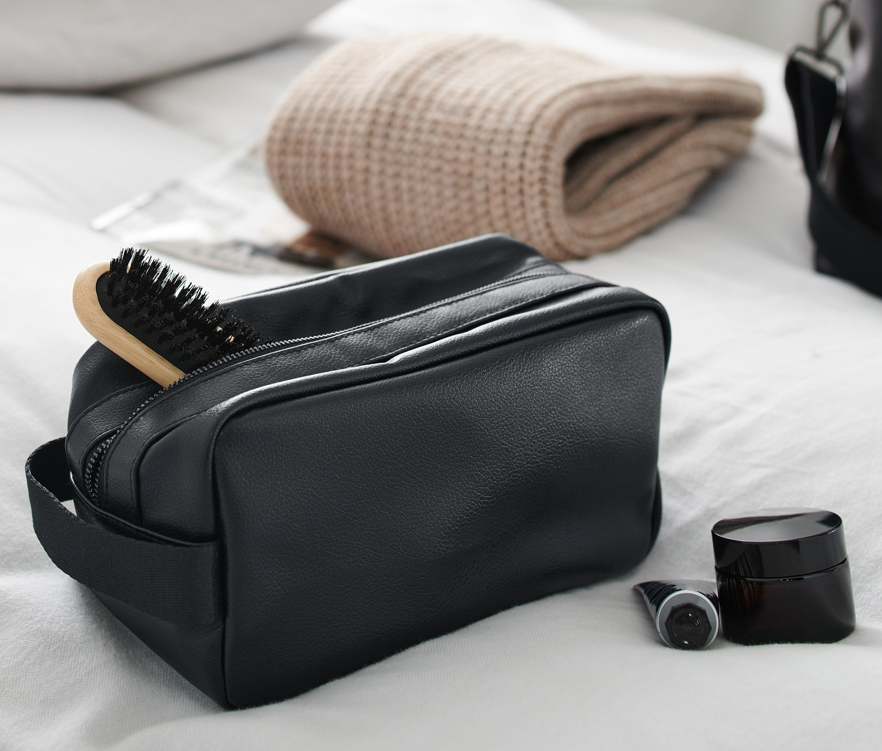 Eine schwarze Kulturtasche mit Haarbürste, Creme und Flasche auf einem weißen Bettlaken.
