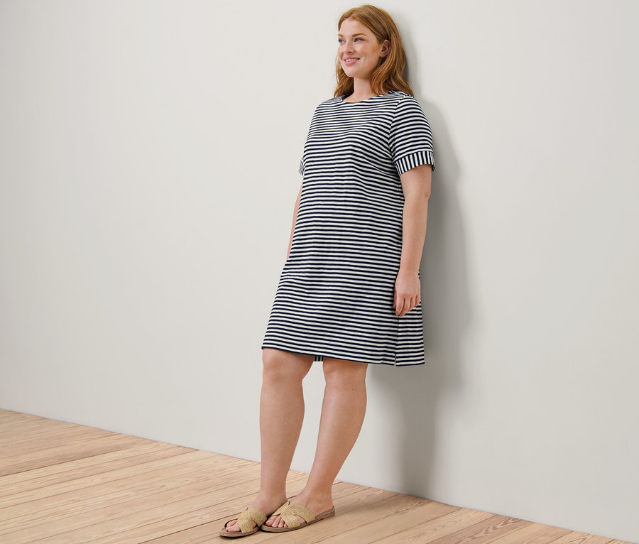 Eine rothaarige Frau lächelt, während sie in sandfarbenen Sandalen und einem gestreiften Jerseykleid an eine Wand gelehnt ist.