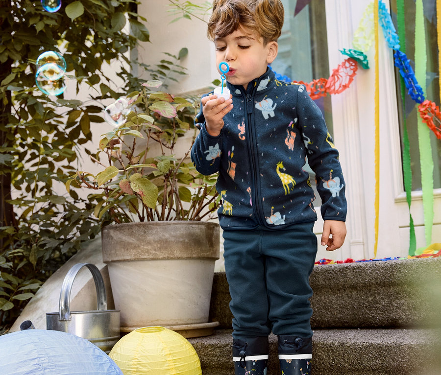 Junge pustet Blasen, trägt eine Kinder-Fleecejacke, Fleecehose und Gummistiefel.