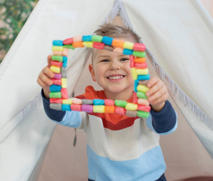 Ein Junge sitzt in einem Tipi und hält ein farbenfrohes Quadrat aus dem PlayMais®-Set in den Händen.