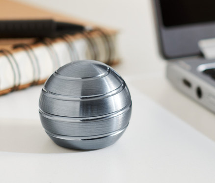 Ein metallischer Mezmoglobe® Spinner liegt auf einem weißen Tisch neben einem Notebook und einem Notizbuch.