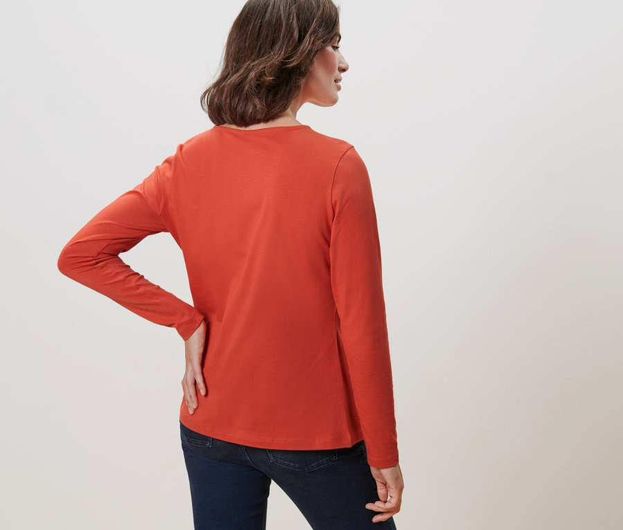 Frau steht mit dem Rücken zur Kamera und trägt ein oranges Langarmshirt.