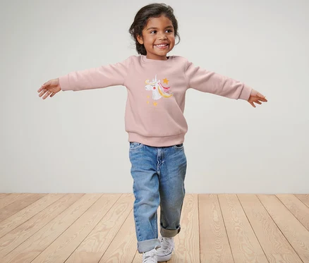 Mädchen mit ausgestreckten Armen trägt einen Kinder-Sweater mit Einhorn, blaue Jeans und Turnschuhe.