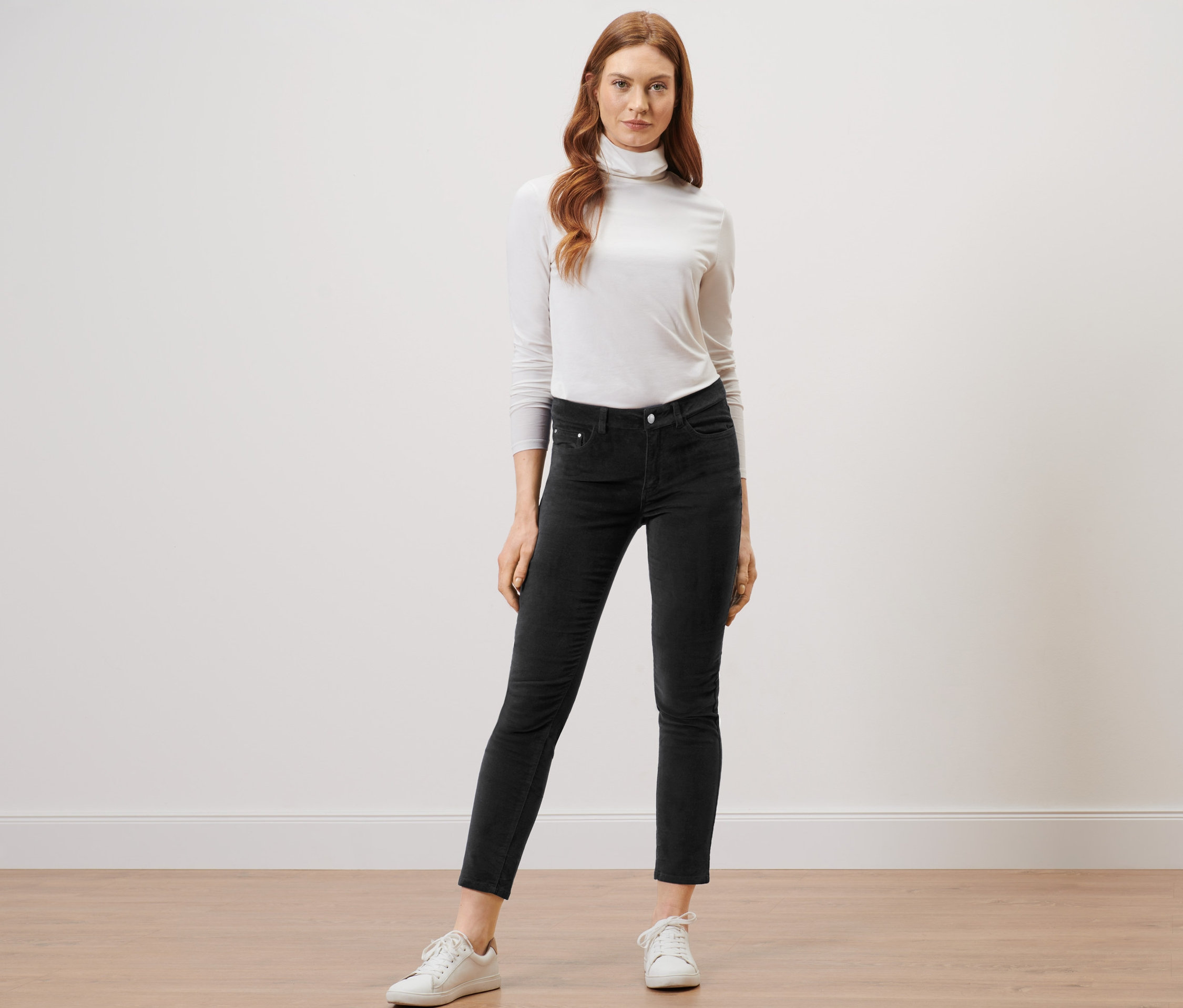 Frau steht auf einem Holzboden und trägt eine schwarze Samthose und einen weißen Rollkragenpullover.