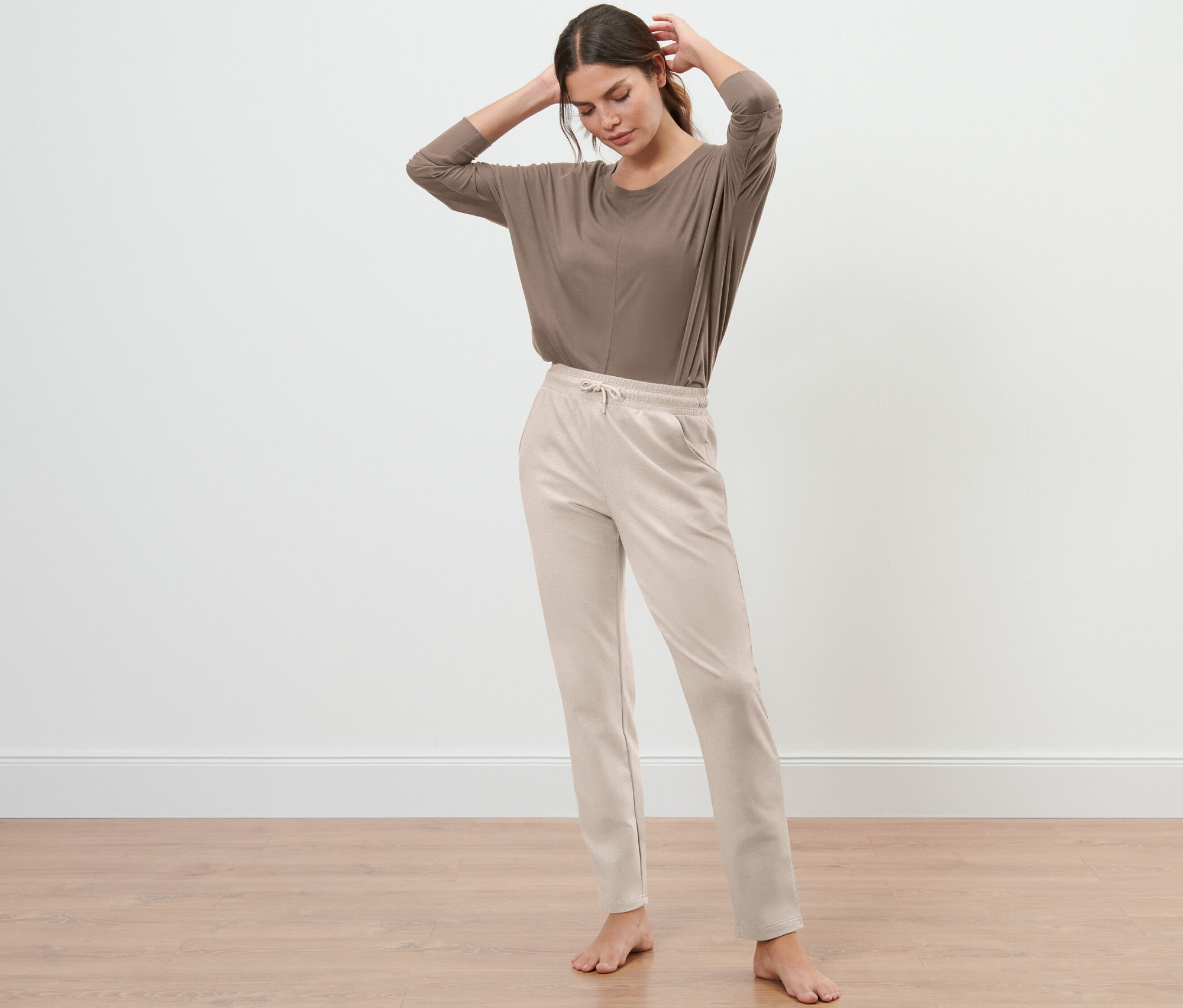 Frau steht mit den Händen am Hinterkopf und trägt eine beige Sweathose.