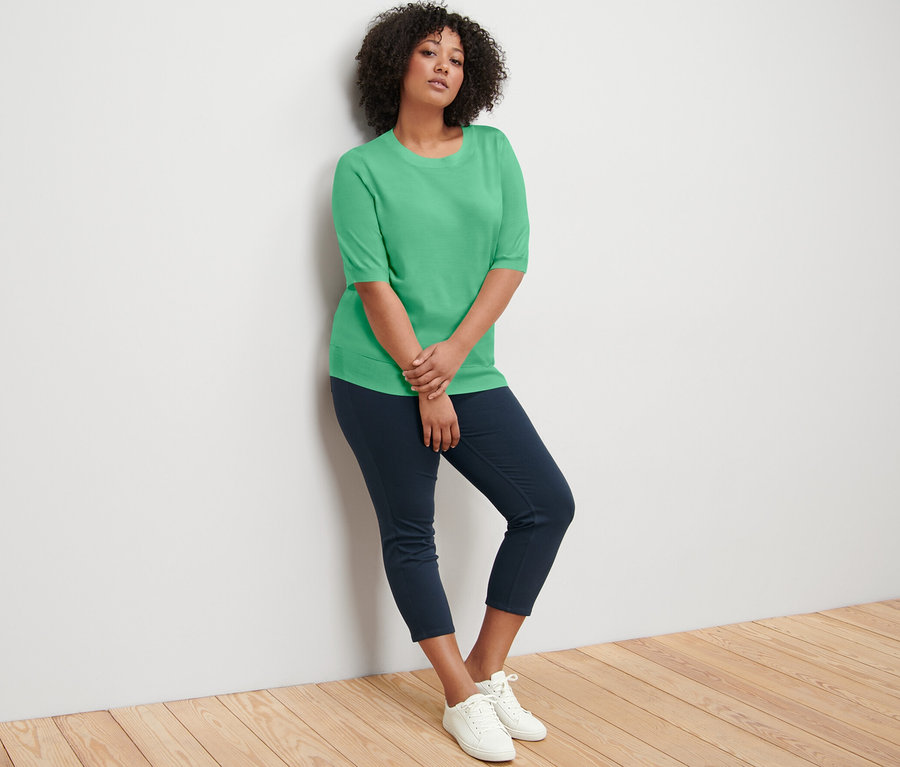 Eine Frau lehnt an einer Wand und trägt einen grünen Feinstrickpullover, dunkelblaue Hosen und weiße Turnschuhe.