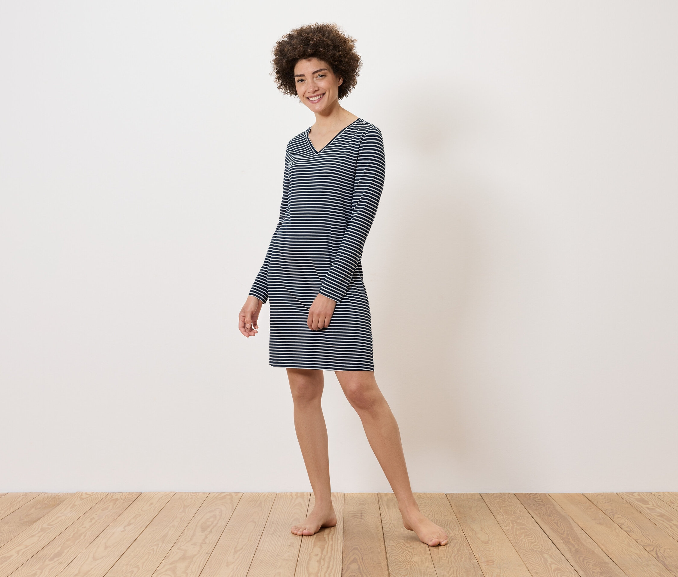 Frau steht barfuß auf Holzboden und trägt ein gestreiftes, langärmeliges Kleid.