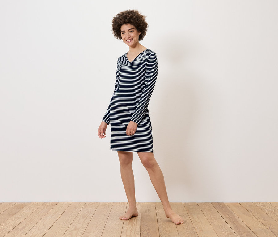 Frau steht barfuß auf Holzboden und trägt ein gestreiftes, langärmeliges Kleid.