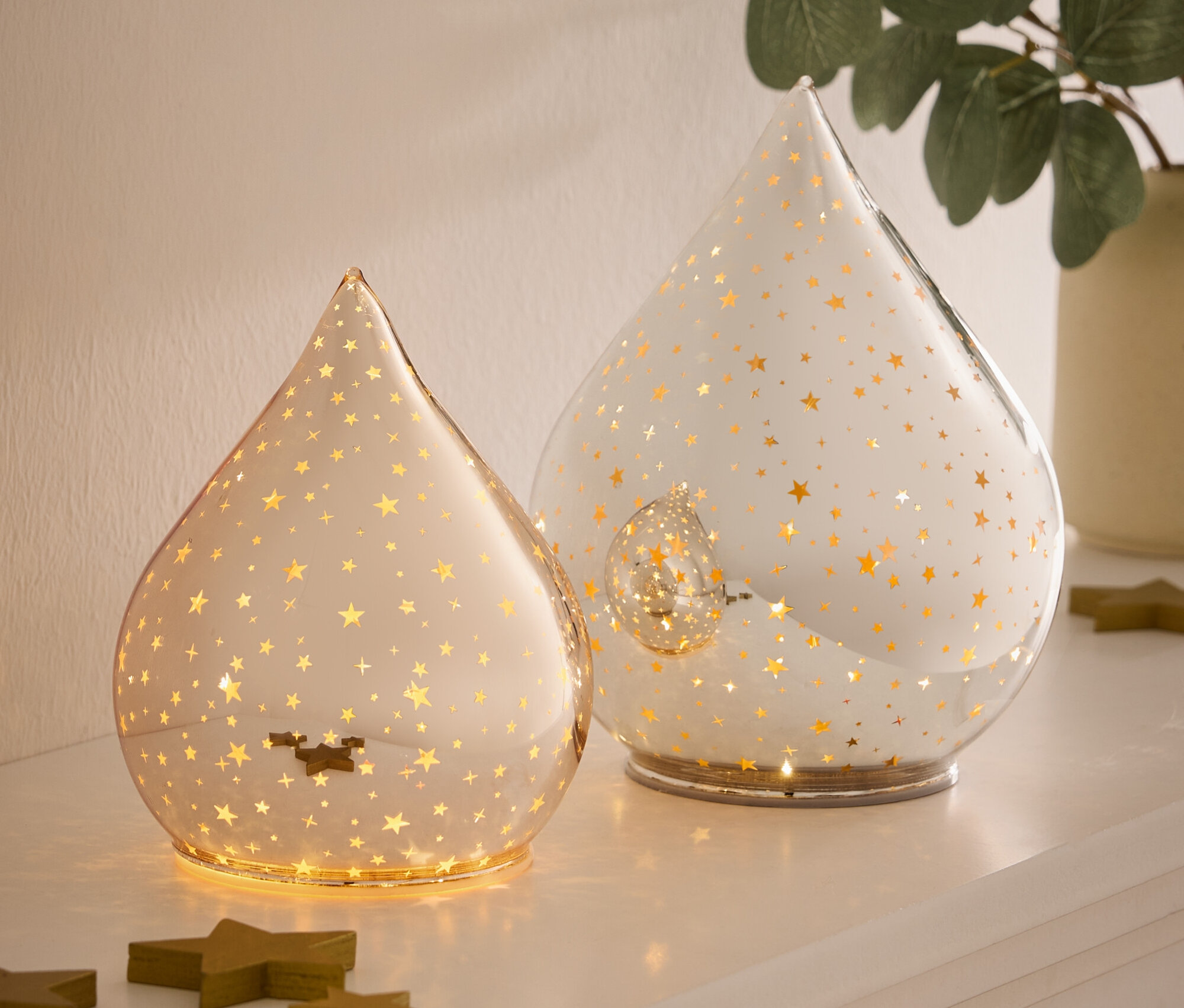 Zwei tropfenförmige Glaslampen mit goldenen Sternen stehen auf einer weißen Oberfläche neben grünen Holzsternen.