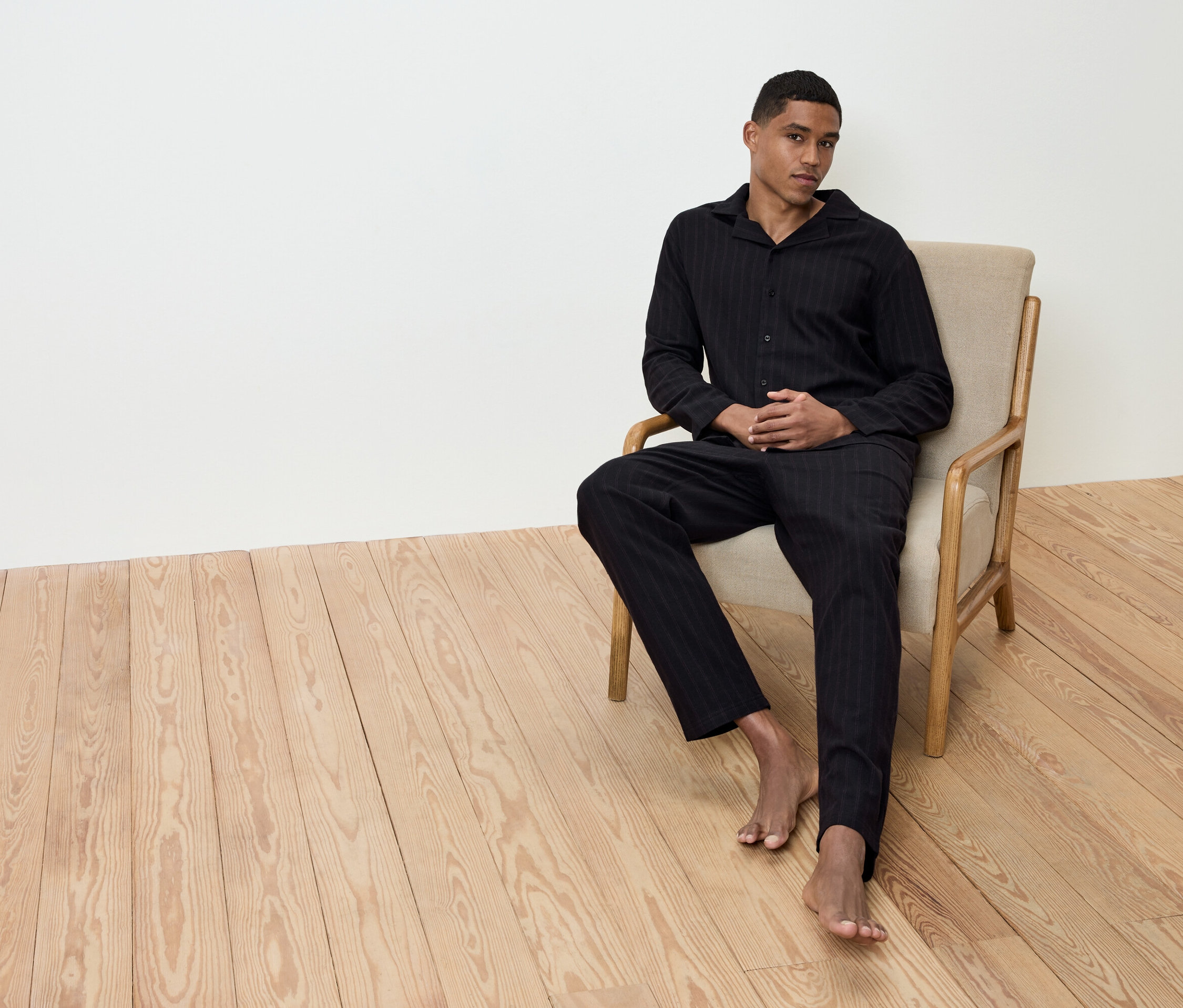 Mann sitzt barfuß in beigem Sessel auf Holzboden, trägt schwarz gestreiften Pyjama.