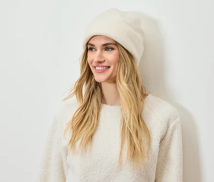 Lächelnde Frau mit blonden Haaren trägt eine cremefarbene Mütze und einen Pullover.