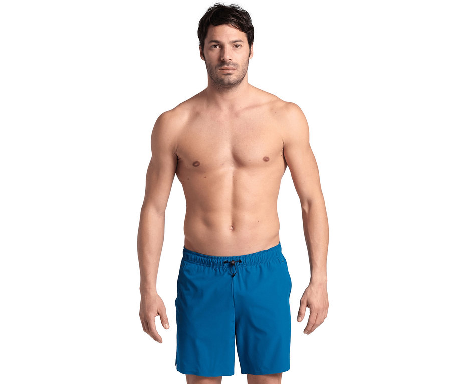 Mann mit braunen Haaren und ohne Hemd posiert vor einem weißen Hintergrund und trägt blaue Badeshorts.