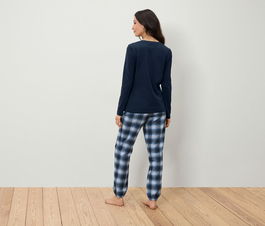 Frau in dunkelblauem Pyjama mit karierter Hose steht mit dem Rücken zur Kamera auf einem Holzboden vor einer weißen Wand.