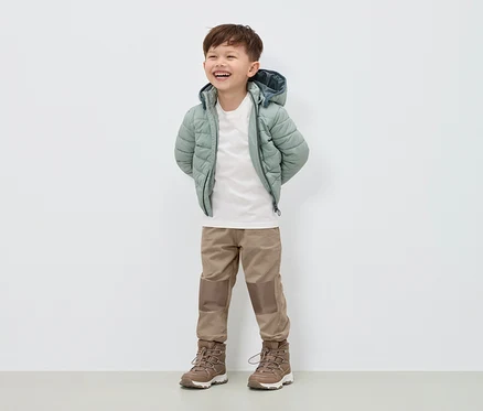 Ein lächelnder Junge steht in einer hellgrünen Daunenjacke, beigen Hosen und braunen Stiefeln.