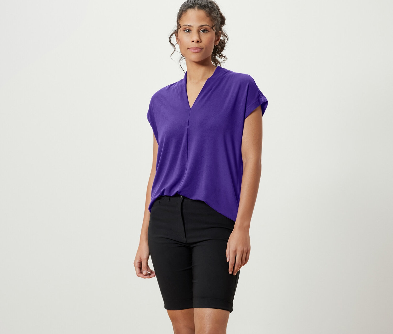 Eine Frau steht und trägt ein lila Blusenshirt mit V-Ausschnitt, schwarze Shorts und weiße Turnschuhe.