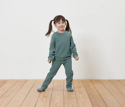 Ein lächelndes kleines Mädchen mit Zöpfen posiert in einem hellgrünen Sweatshirt- und Jogginghosen-Set auf einem Holzboden vor einer weißen Wand.