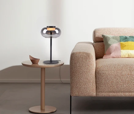 Graue Lampe steht auf rundem Tisch neben beigem Sofa mit buntem Kissen.