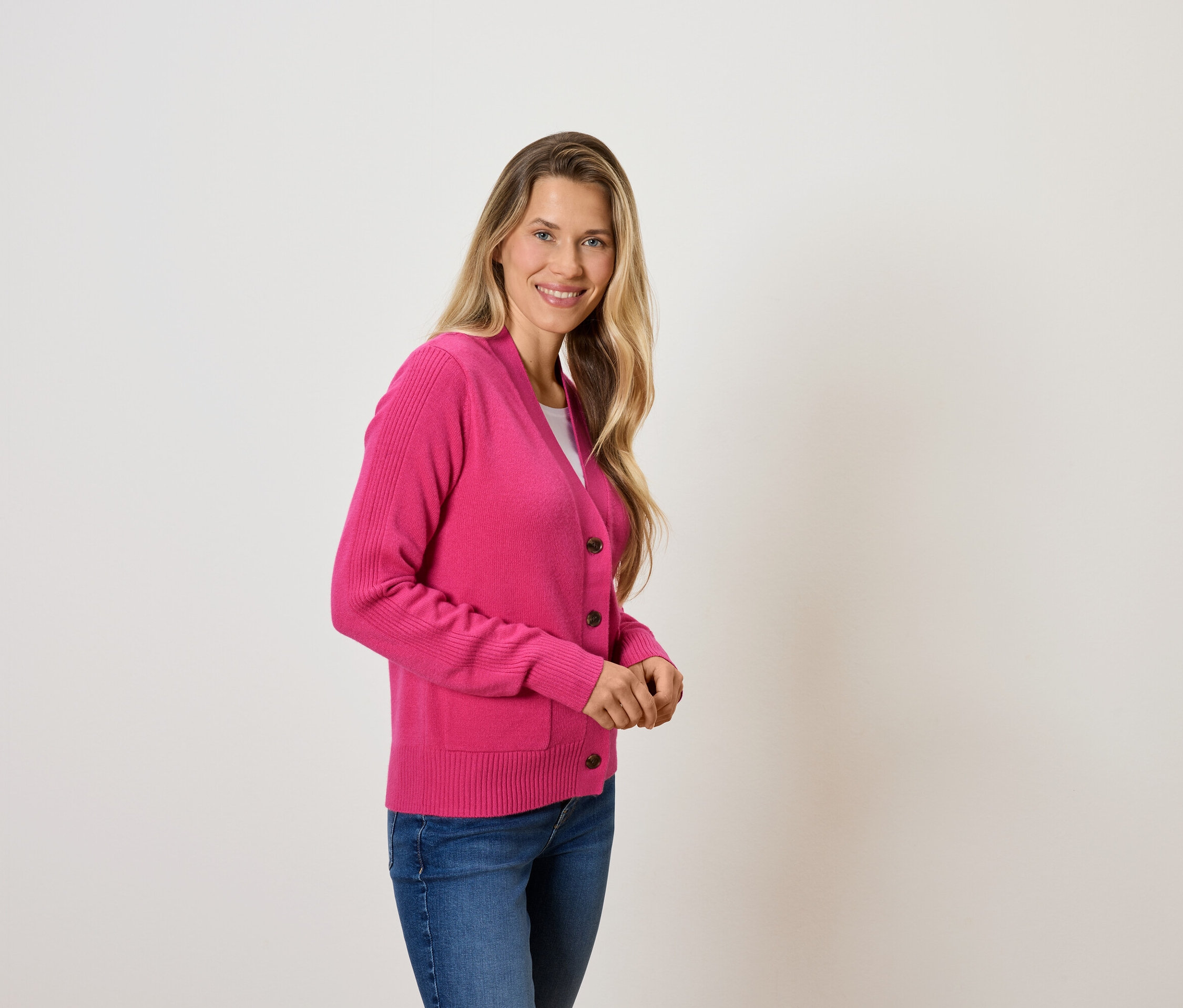 Frau mit blonden Haaren posiert in einem pinkfarbenen Cardigan und blauen Jeans.