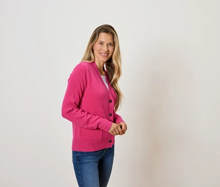 Frau mit blonden Haaren posiert in einem pinkfarbenen Cardigan und blauen Jeans.