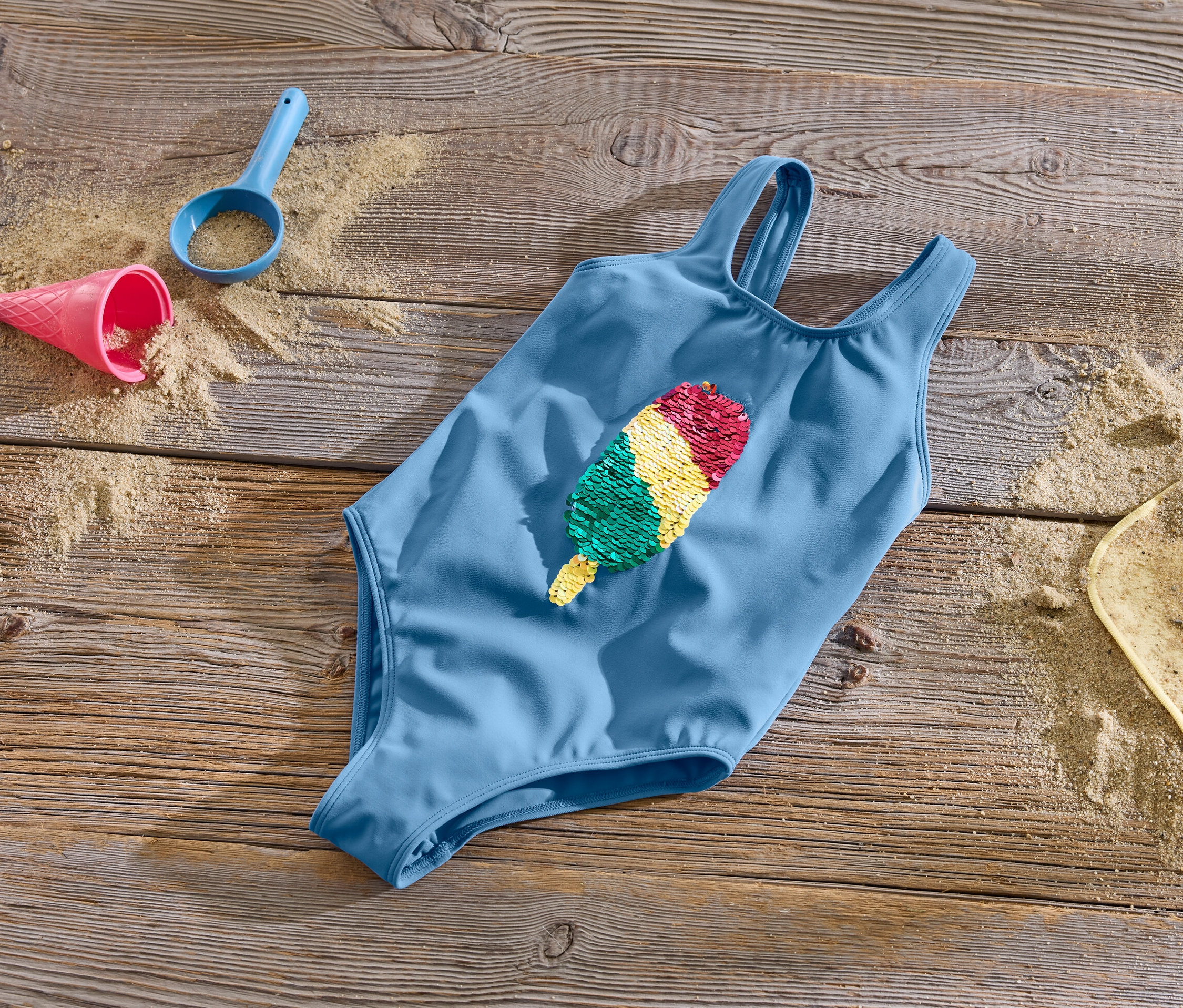 Blauer Kinder-Badeanzug mit Wendepailletten liegt auf einer Holzoberfläche mit Sand und Spielzeug.