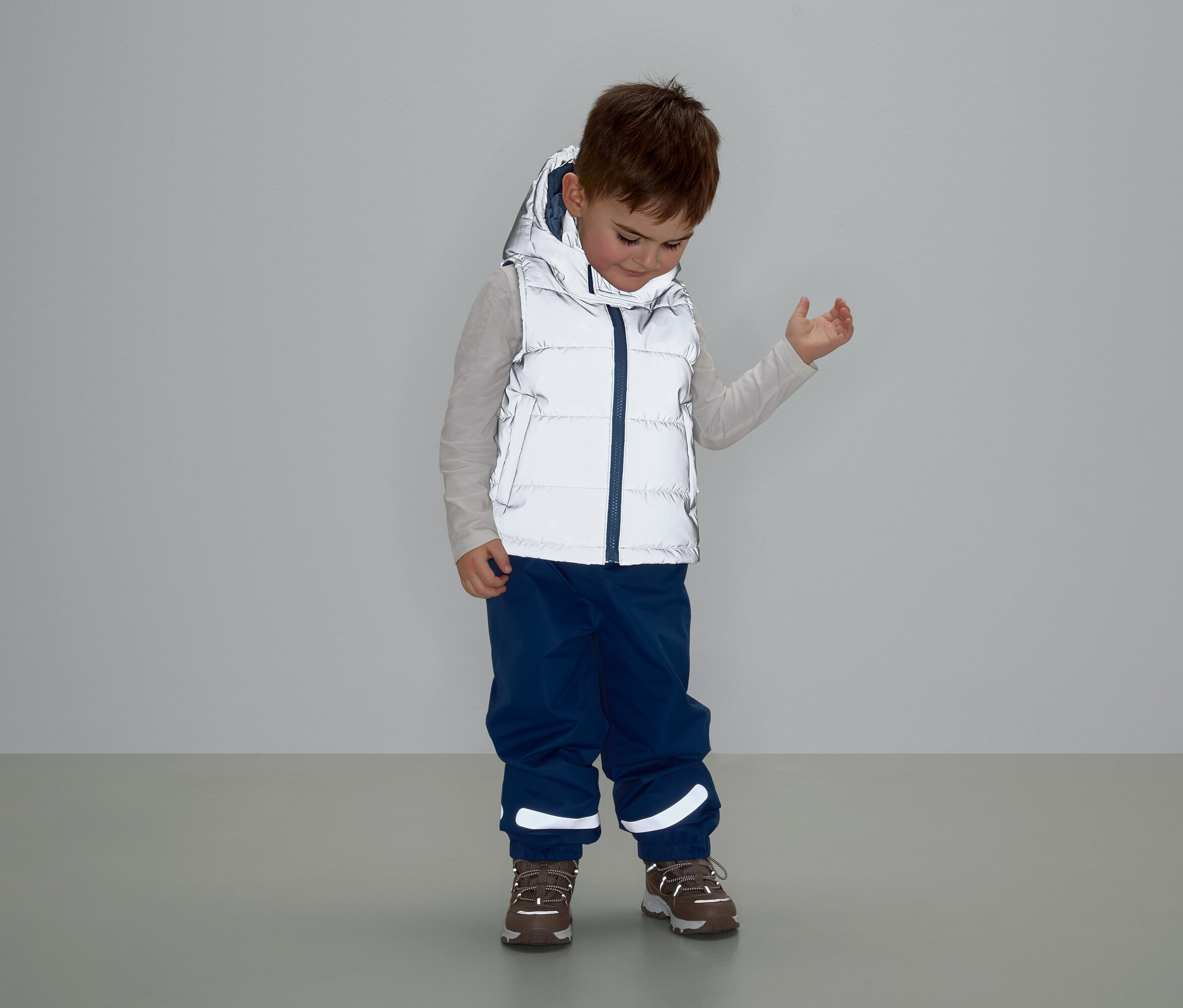 Junge mit grauer Weste mit Kapuze, blauer Hose und braunen Schuhen steht und schaut nach unten.