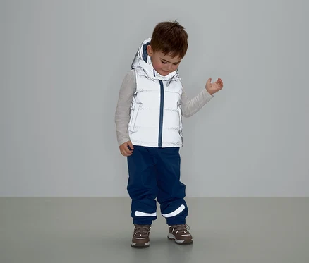 Junge mit grauer Weste mit Kapuze, blauer Hose und braunen Schuhen steht und schaut nach unten.