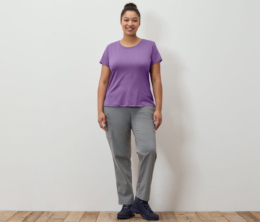 Eine Frau steht und trägt eine graue, packbare Outdoorhose und ein Stretch-Funktionsshirt.