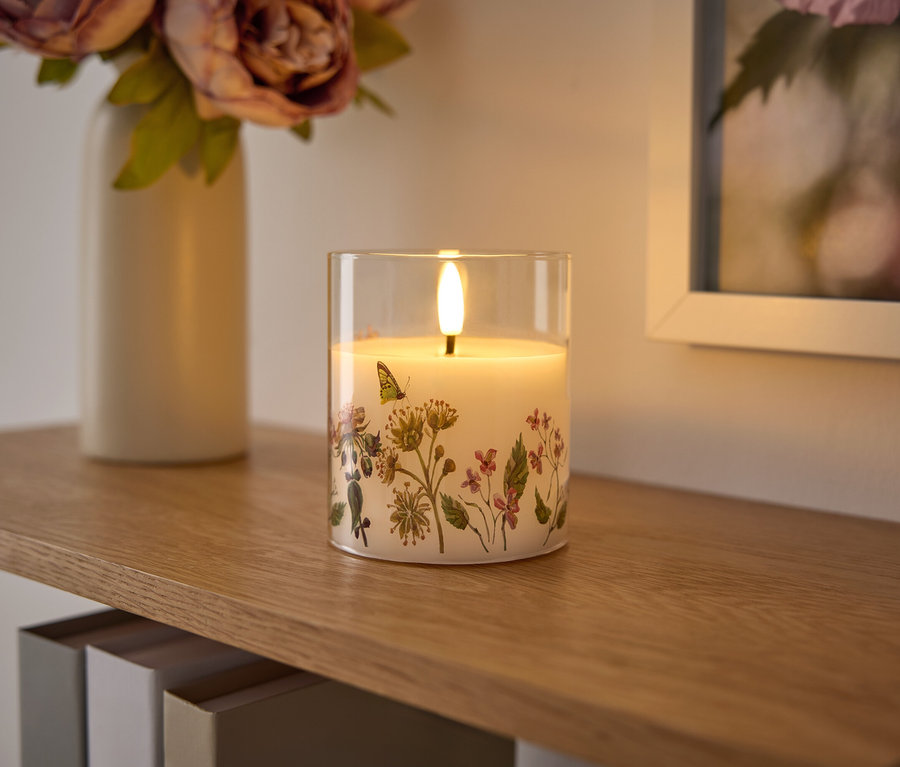 Auf einem Holzregal stehen eine Glas-Kerze mit Blumenmuster, eine Blumenvase und ein gerahmtes Bild.