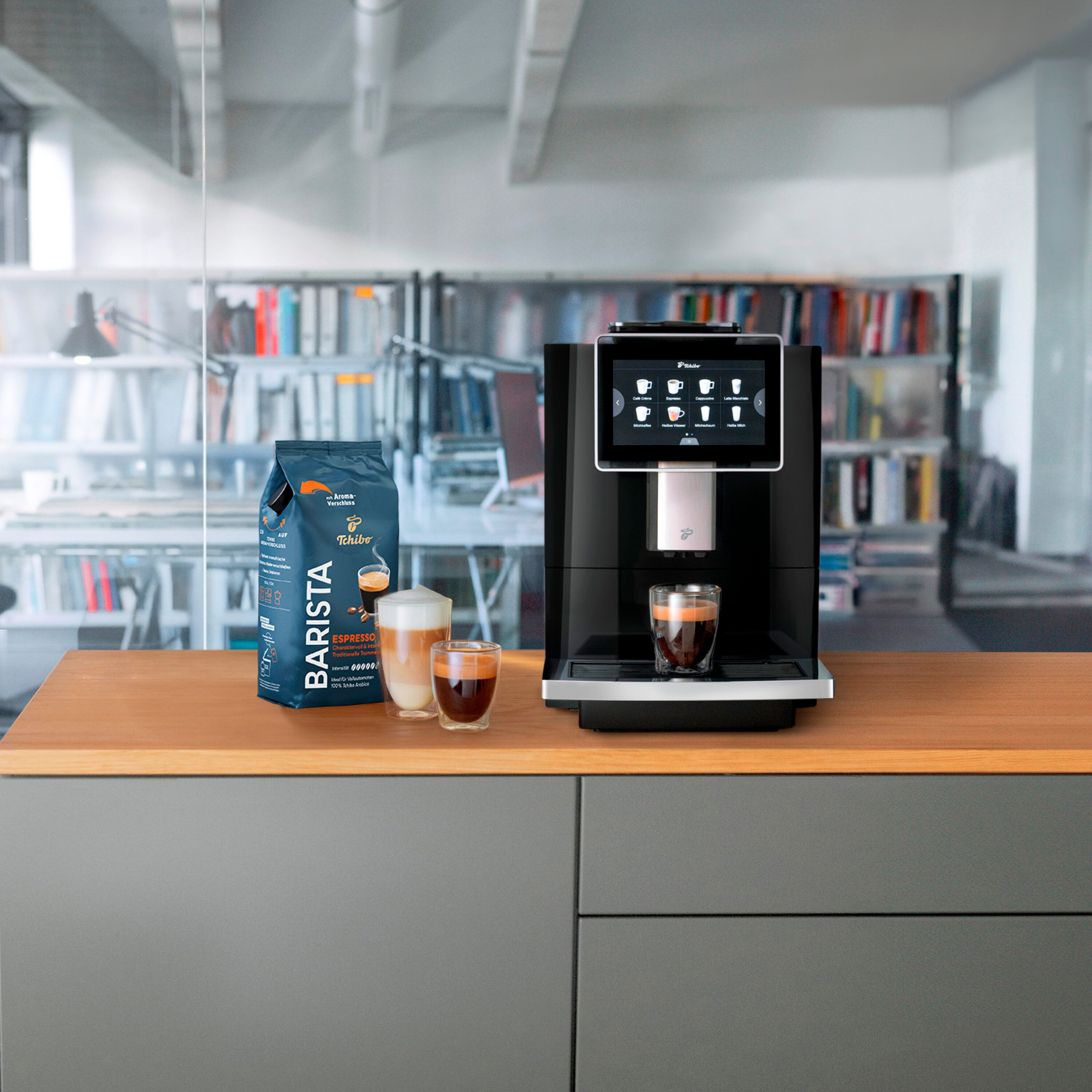 Ein Kaffeevollautomat steht neben einer Packung Barista-Kaffee und zwei Gläsern Kaffee.