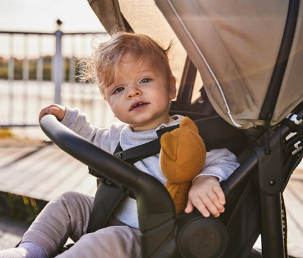 Ein blondes Kind sitzt mit einem braunen Stofftier in einem Kinderwagen.