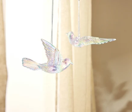 Zwei schimmernde Vogelornamente hängen vor einem beigen Vorhang.