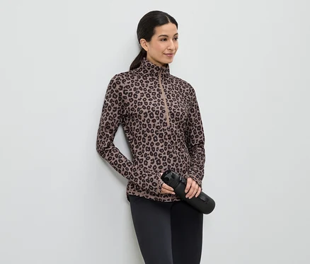Frau mit braunem Leopard-Print-Oberteil, schwarzen Leggings und schwarzer Wasserflasche, die an einer weißen Wand lehnt.
