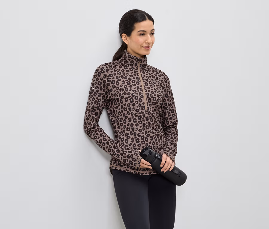 Frau mit braunem Leopard-Print-Oberteil, schwarzen Leggings und schwarzer Wasserflasche, die an einer weißen Wand lehnt.