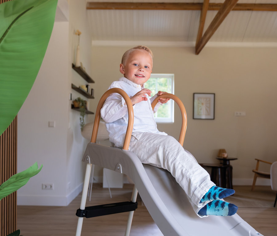 Ein lächelnder Junge in weißem Hemd und Hose sitzt auf einer grauen Rutsche mit Holzgriffen in einem hellen Wohnzimmer.