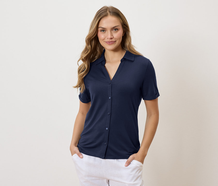 Frau mit langen, gewellten, blonden Haaren, die ein marineblaues Hemd und eine weiße Hose trägt und die Hände in den Hosentaschen hat.