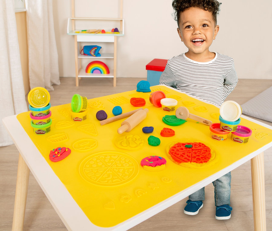 Ein lächelnder Junge steht an einem Tisch mit gelber Spielmatte, Play-Doh-Dosen und Werkzeugen zum Formen von Knete.