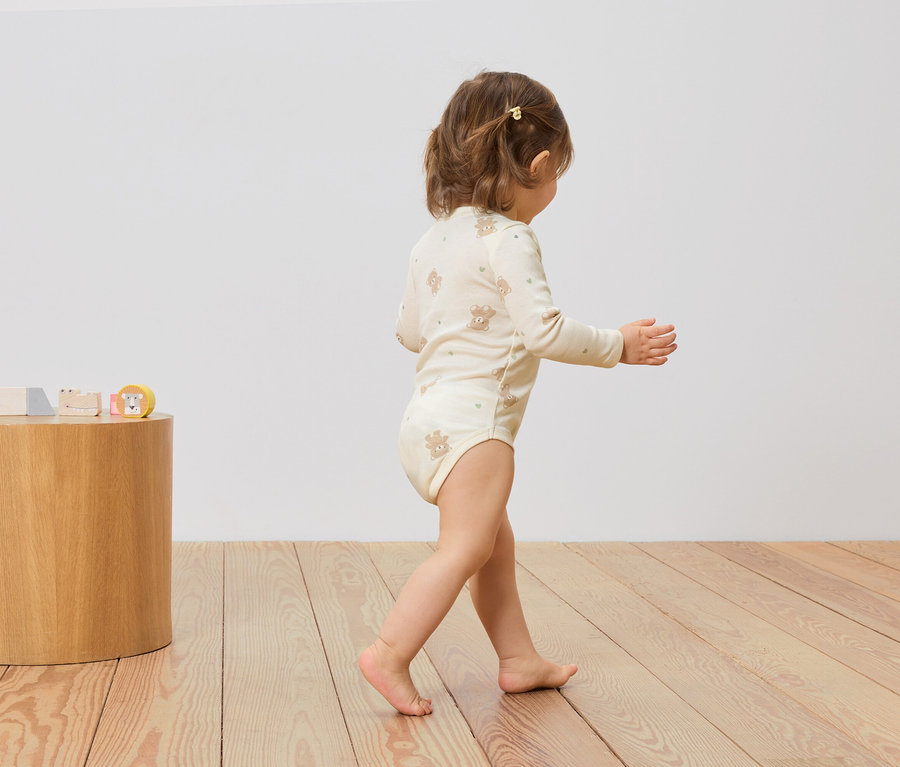 Kleinkind in einem beigen Body mit Bärendruck geht auf einem Holzboden. Auf einem Holzhocker stehen Holzspielzeuge.