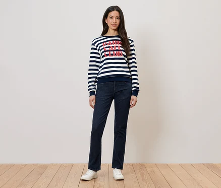 Frau in gestreiftem Pullover mit Aufdruck und dunkler Jeans, dazu weiße Schuhe.