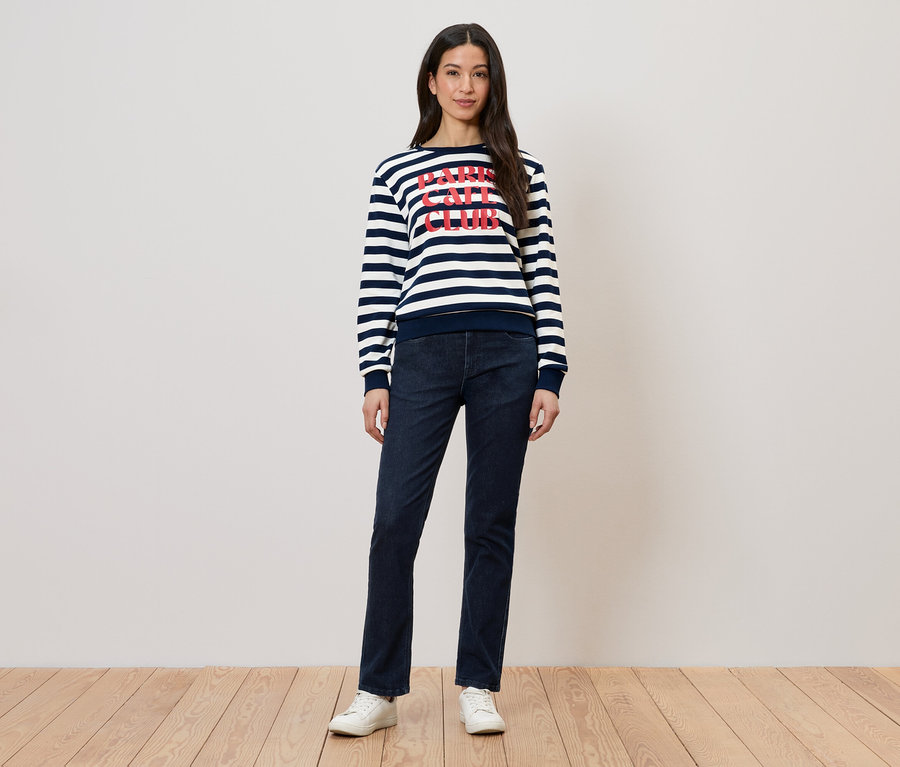 Frau in gestreiftem Pullover mit Aufdruck und dunkler Jeans, dazu weiße Schuhe.