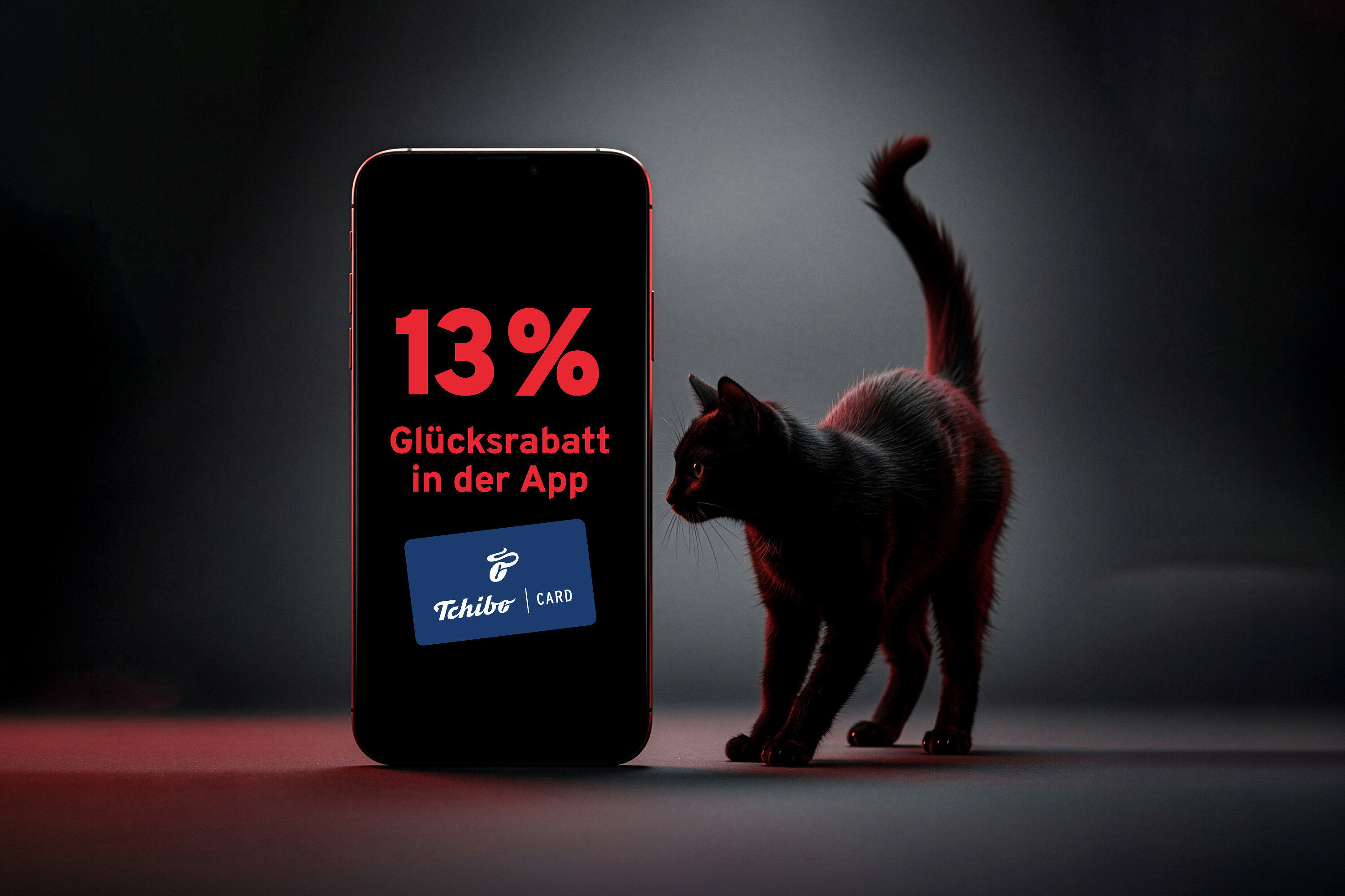 Schwarze Katze steht neben einem Smartphone mit der Aufschrift 13% Glücksrabatt in der App und einer Tchibo Card.