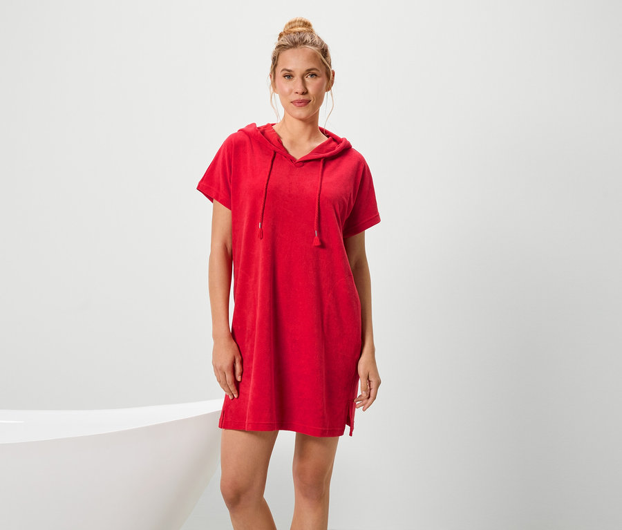 Eine Frau steht neben einer Badewanne und trägt ein rotes, kurzärmeliges Kapuzenkleid.