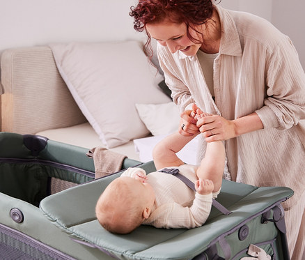 Lächelnde Frau hält die Füße eines Babys, das auf einem Wickeltisch in einem Kinderbett liegt.
