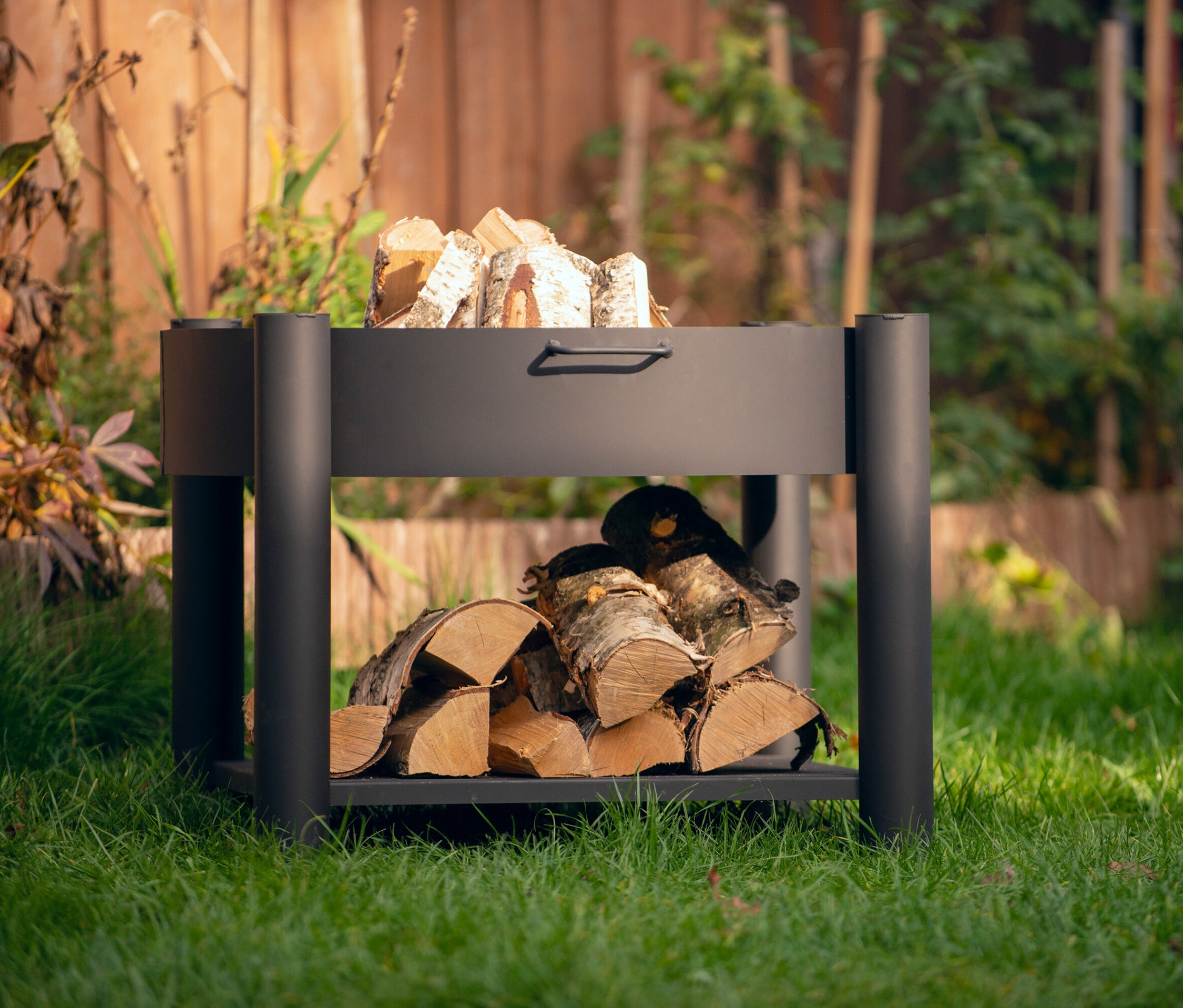 Ein schwarzer Holzkorb steht auf einem Rasen, gefüllt mit Holzscheiten.