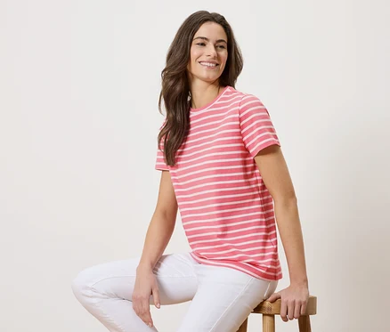Eine Frau sitzt auf einem Hocker und trägt ein rosa-weiß gestreiftes T-Shirt und weiße Jeans.