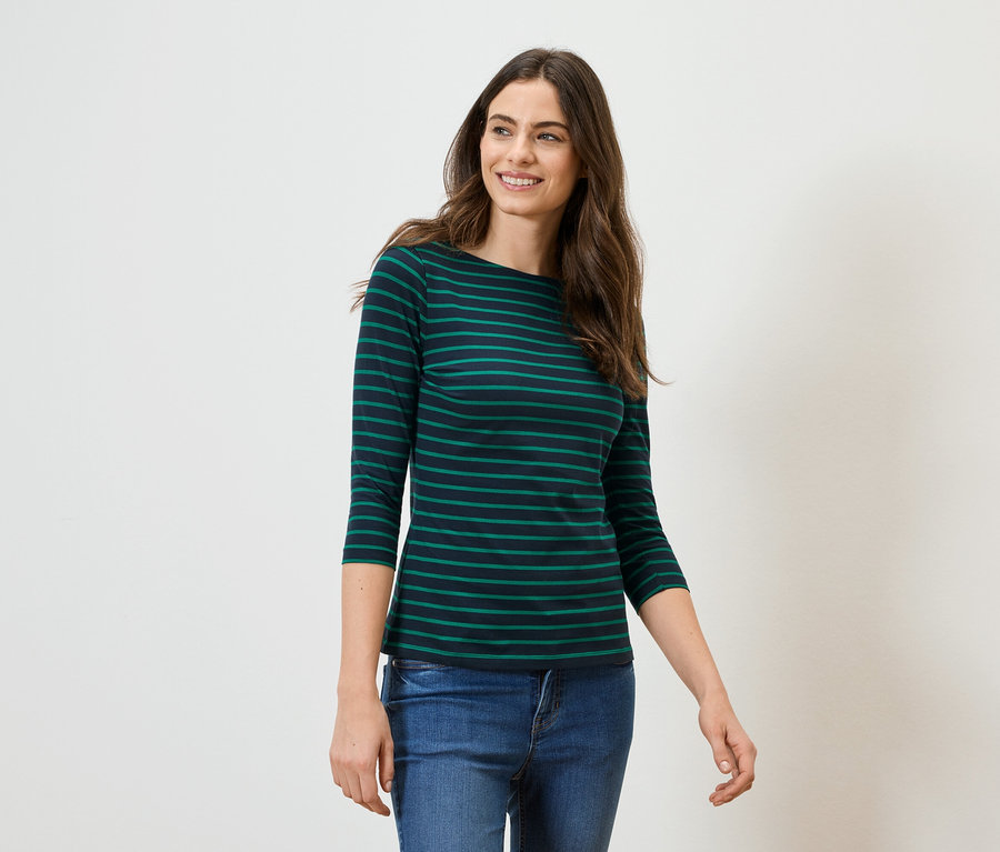 Frau mit langen braunen Haaren trägt ein grün-schwarz gestreiftes Langarmshirt und Jeans vor einem weißen Hintergrund.