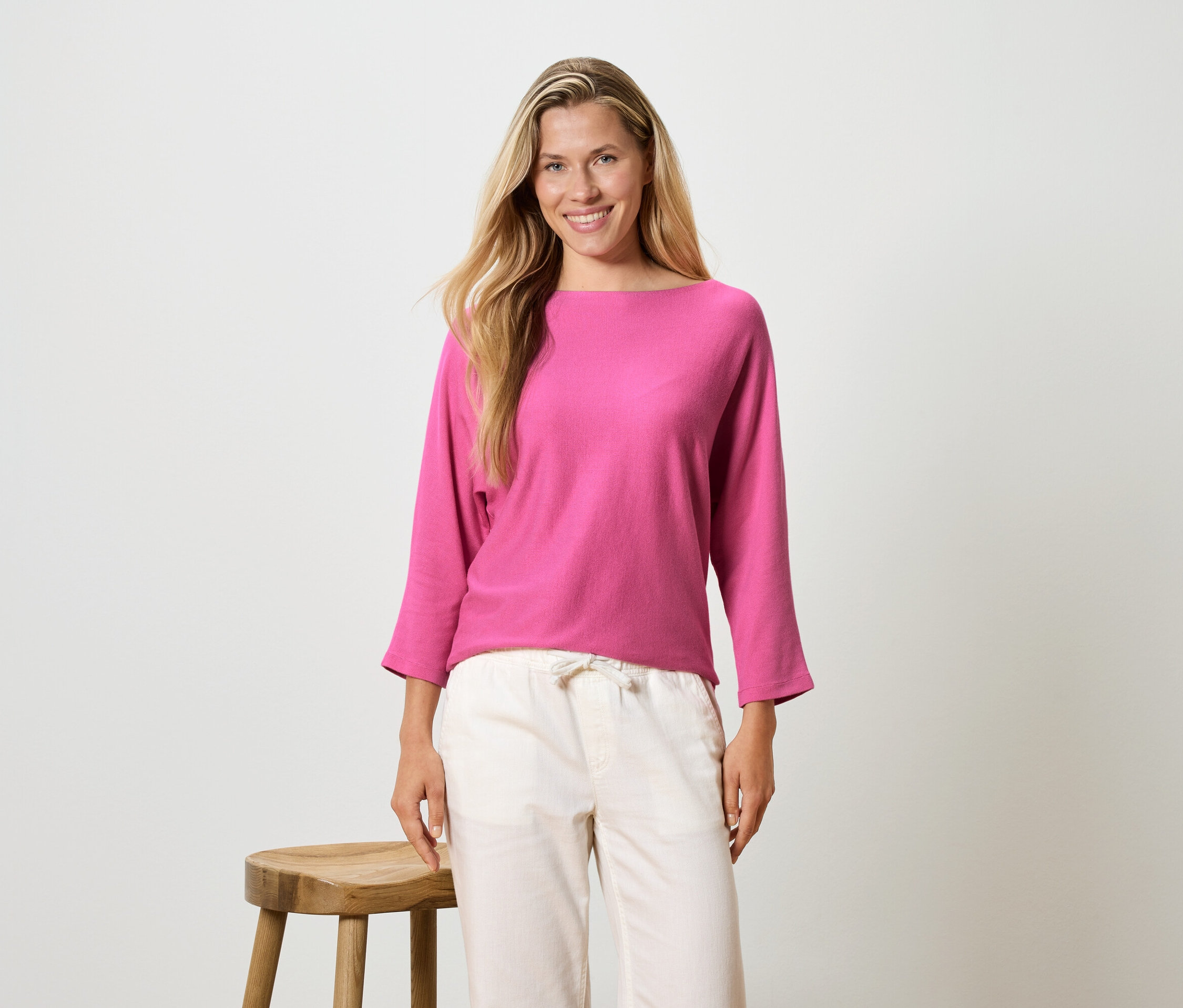 Frau mit langen blonden Haaren trägt ein pinkfarbenes Langarmshirt und eine helle Hose und steht neben einem Hocker.