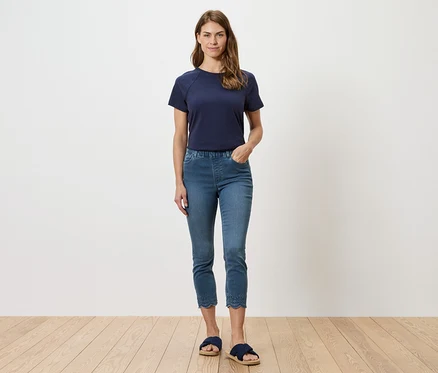 Frau in marineblauem T-Shirt und blauen Jeans mit Lochstickerei am Saum, posiert vor einer weißen Wand.